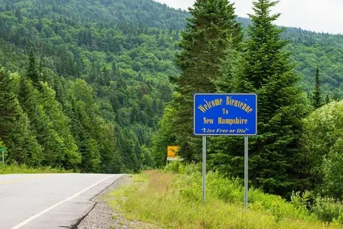 A roadside sign reading "Welcome to New Hampshire" with a lush evergreen landscape behind it.